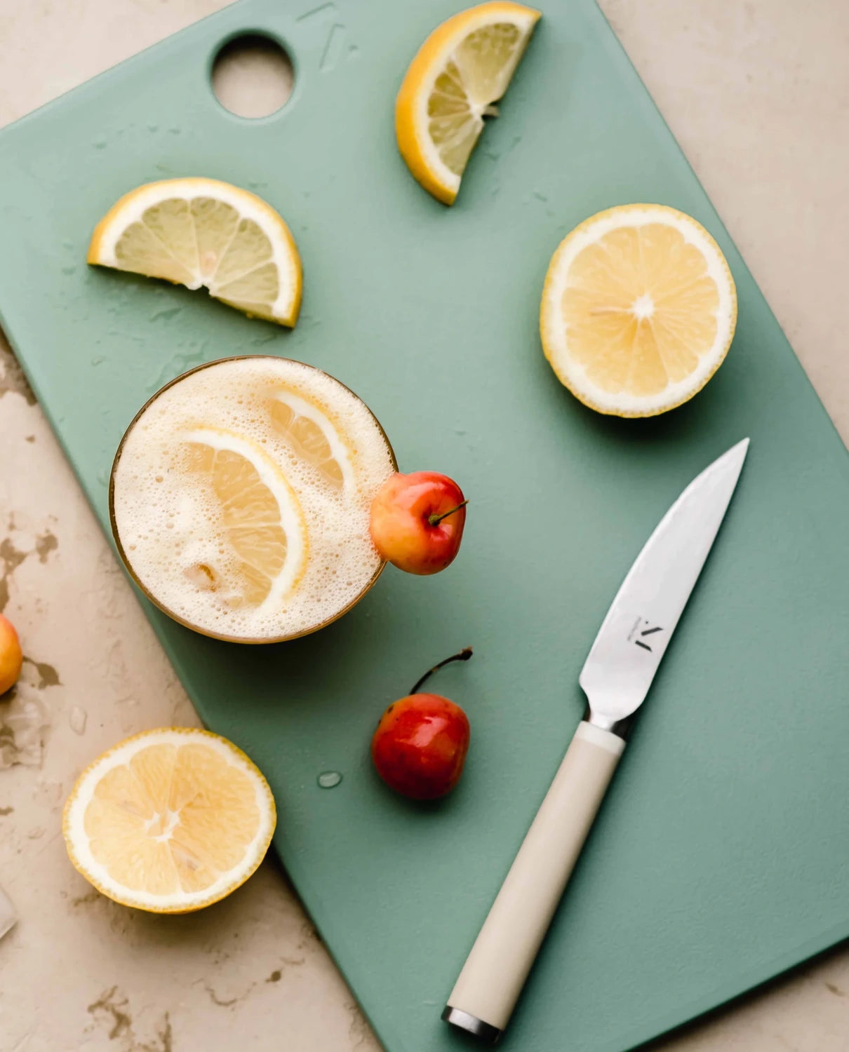 Sage green cutting board with lemon slices, whole lemons, and white handled knife on marble countertop