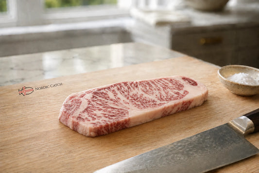 A raw, highly marbled wagyu NY strip steak rests on a wooden cutting board, with a chef's knife and a small bowl of sea salt nearby in a bright kitchen setting. The 'NORDIC CATCH' brand logo is visible on the left side of the board.