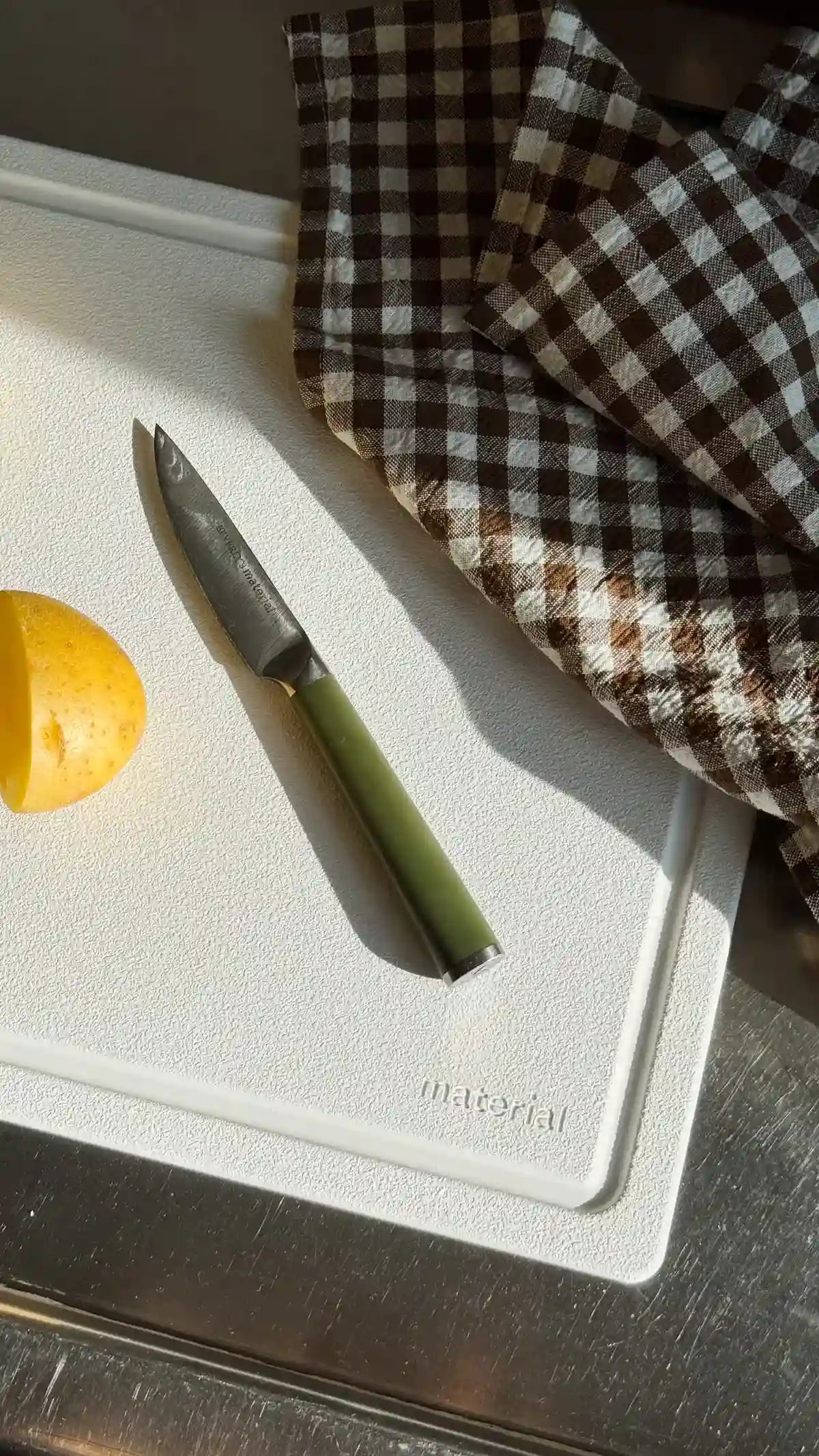 Non-toxic cutting board with chef knife and halved lemon on white surface with brown checkered kitchen towel