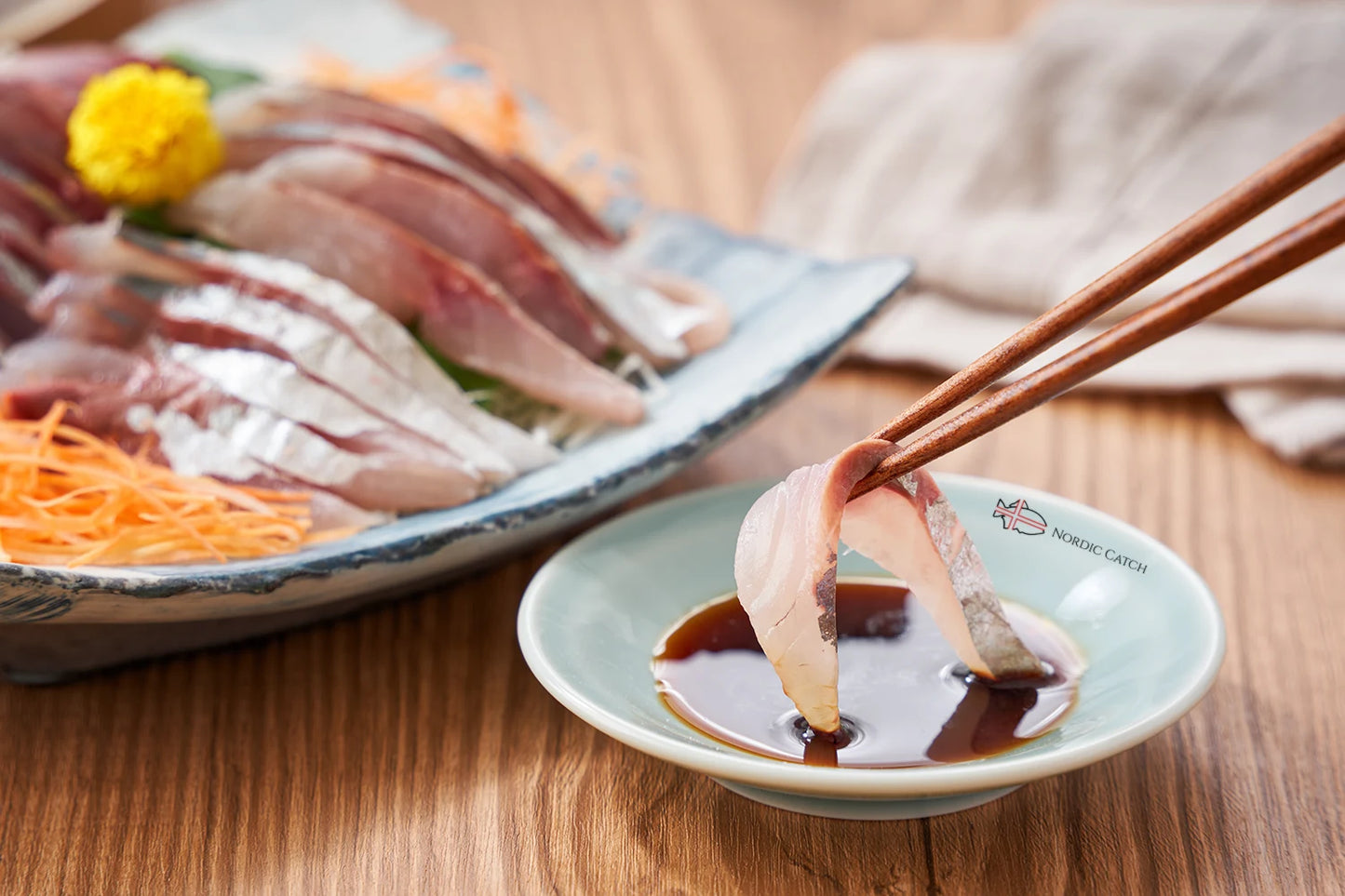 Horse mackerel sashimi slice held with wooden chopsticks over Nordic Catch branded dipping sauce on wooden surface with assorted sashimi platter in background