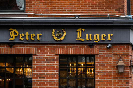 Peter Luger Steak House storefront with gold lettering, established 1887 laurel wreath logo, and lit windows on red brick exterior