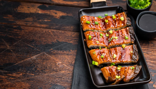 Best wild-caught fish unagi eel fillets sliced and glazed in teriyaki sauce, topped with sesame seeds and scallions on a black rectangular plate