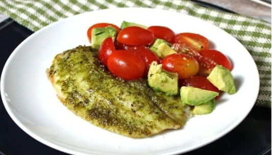 Fresh fish fillet coated in pesto, served with cherry tomatoes and diced avocado on a white plate