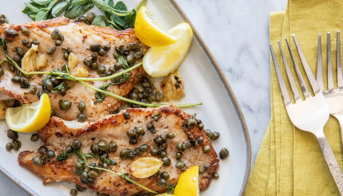 Healthy seafood recipes featuring pan-seared fish fillets topped with capers, garlic, fresh herbs, and lemon wedges on a white plate