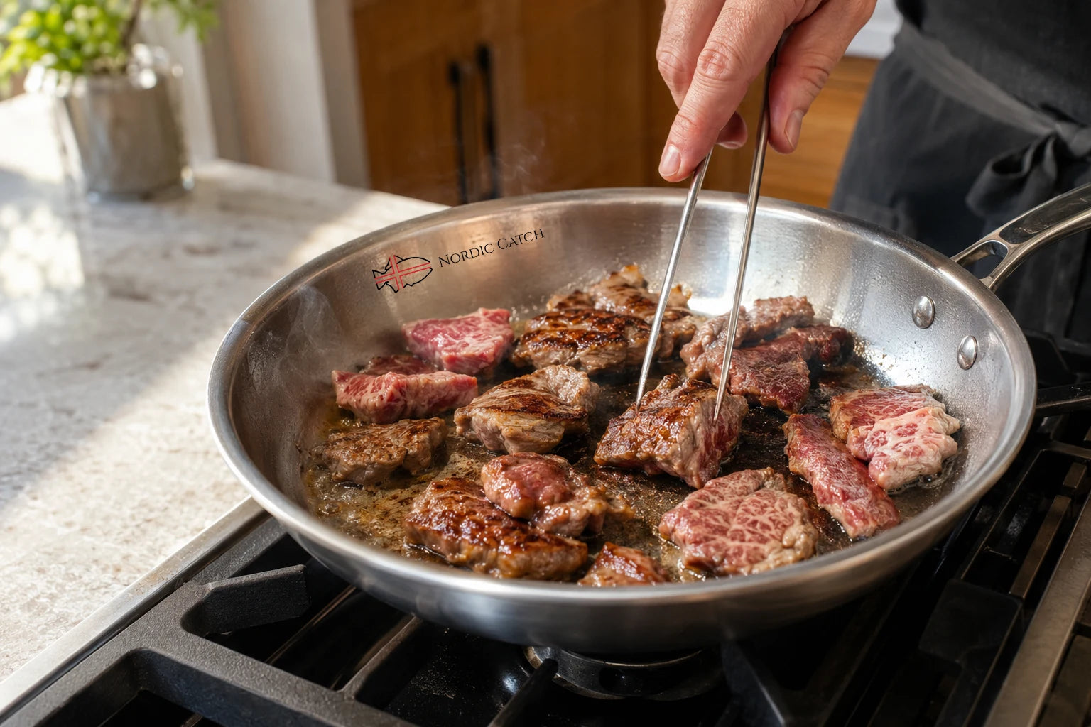 A stainless steel skillet on a stovetop with wagyu steak pieces cooking in oil, held by a person using metal tongs. The skillet has the 'Noetic Cookware' brand name printed inside near the rim.