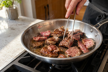 A stainless steel skillet on a stovetop with wagyu steak pieces cooking in oil, held by a person using metal tongs. The skillet has the 'Noetic Cookware' brand name printed inside near the rim.