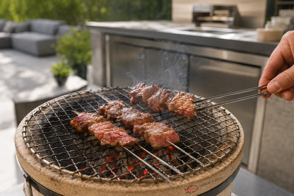 A close-up of skewered wagyu meat grilling on a round charcoal grill outdoors, with a hand using metal tongs to turn the skewers. Smoke rises from the grill, and a modern outdoor kitchen counter is blurred in the background.