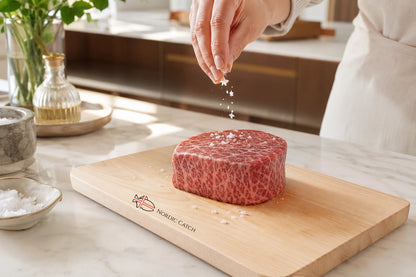 A raw marbled A5 Filet Mignon cut rests on a wooden cutting board with the brand name 'Nordic Caviar' printed on it, while a person's hand sprinkles salt over the meat in a bright kitchen setting.