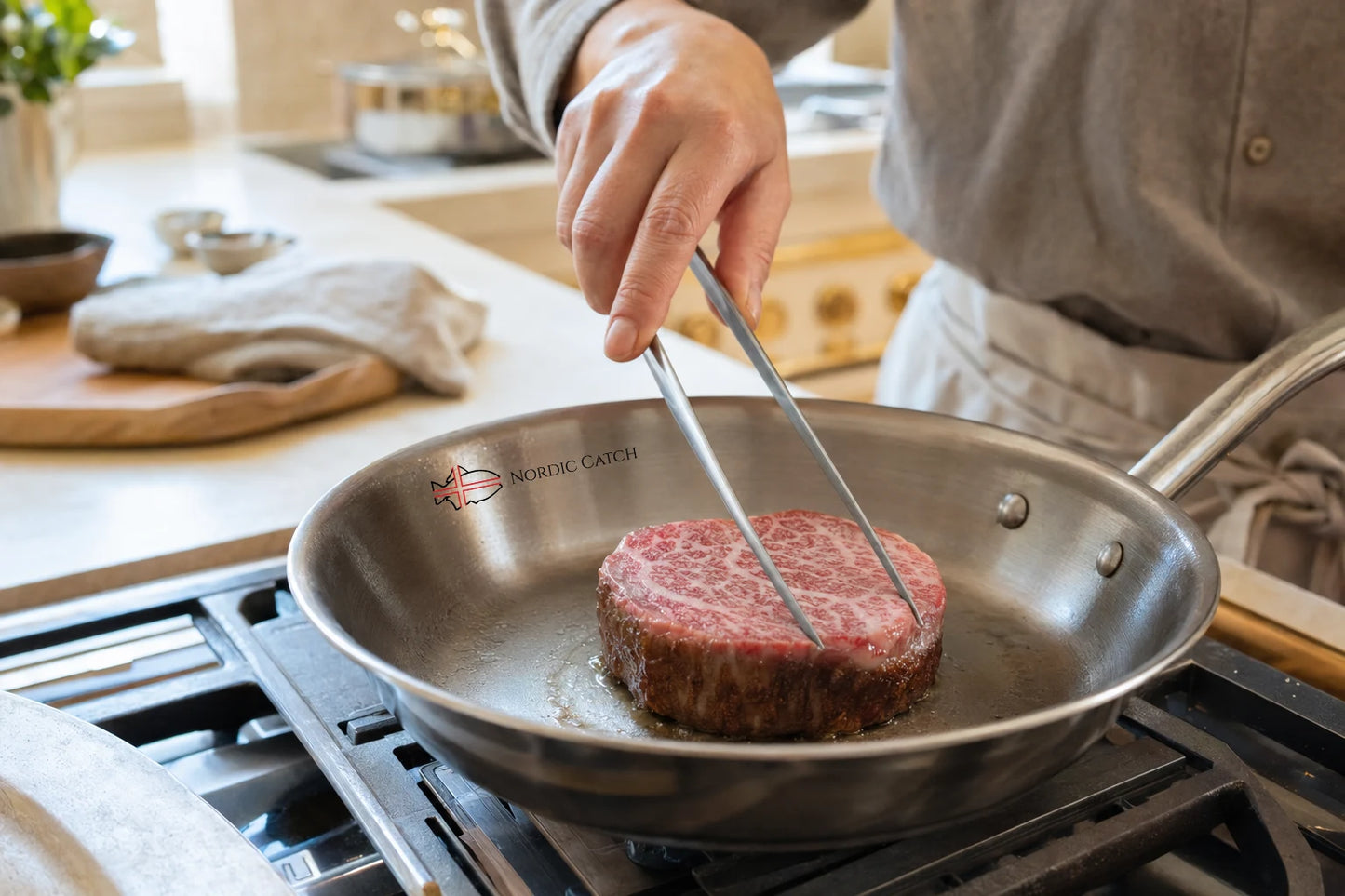 A stainless steel skillet on a stovetop with a raw filet mignon cooking in oil, and a person using metal tongs to lift the steak. The skillet has a visible 'Nordic Catch' logo.