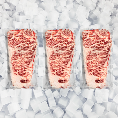 Three raw marbled beef NY strip steaks in clear plastic packaging, arranged side by side on a bed of white ice cubes.