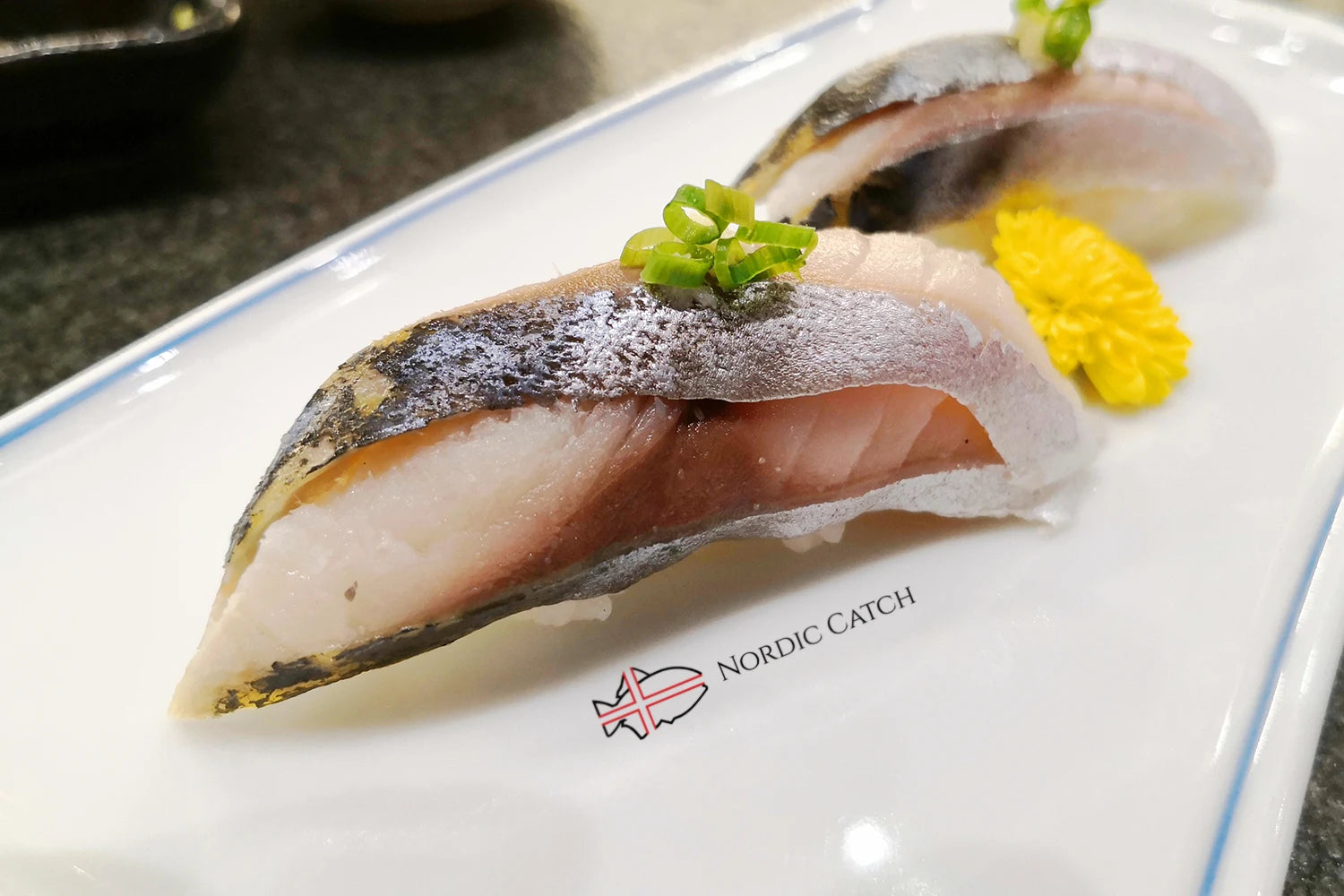 Horse mackerel nigiri sushi with skin charred on top, scallion garnish, and yellow edible flower on Nordic Catch branded white plate