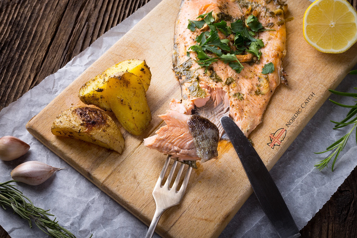 Wild-caught steelhead trout fillet with herb crust and roasted potatoes on Nordic Catch branded wooden board with lemon, fresh rosemary, garlic, and metal utensils