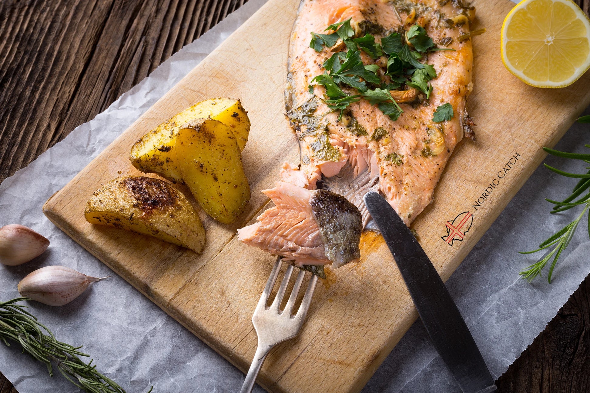 Wild-caught steelhead trout fillet with herb crust and roasted potatoes on Nordic Catch branded wooden board with lemon, fresh rosemary, garlic, and metal utensils