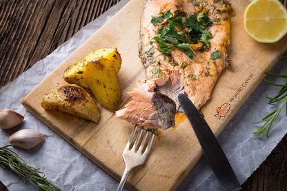 Wild-caught steelhead trout fillet with herb crust and roasted potatoes on Nordic Catch branded wooden board with lemon, fresh rosemary, garlic, and metal utensils
