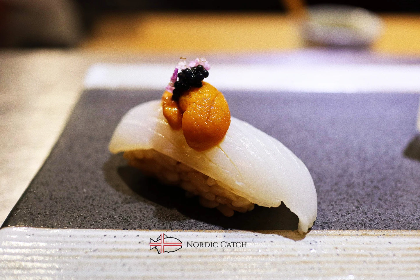 Cuttlefish nigiri sushi topped with uni sea urchin roe, black caviar, and edible flowers on black slate with Nordic Catch branding