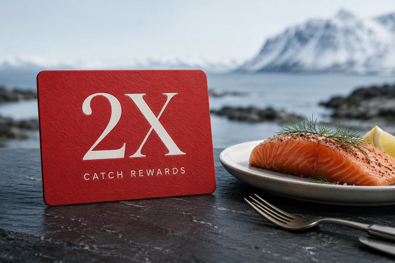 A red Catch Rewards card with large white '2X' text stands on a dark outdoor surface beside a plate of salmon, dill, and lemon, with snowy mountains and water in the background.