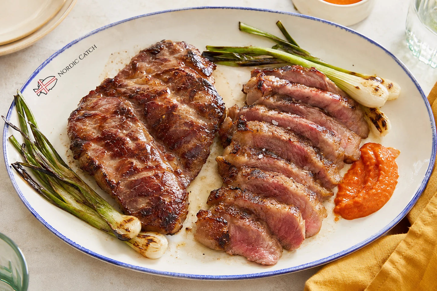 A plated Iberico Pluma steak dish with one seared whole cut and several sliced pieces showing pink medium-rare interiors, served with green onions and a small orange sauce on a white oval plate. The plate is branded Nordic Catch.