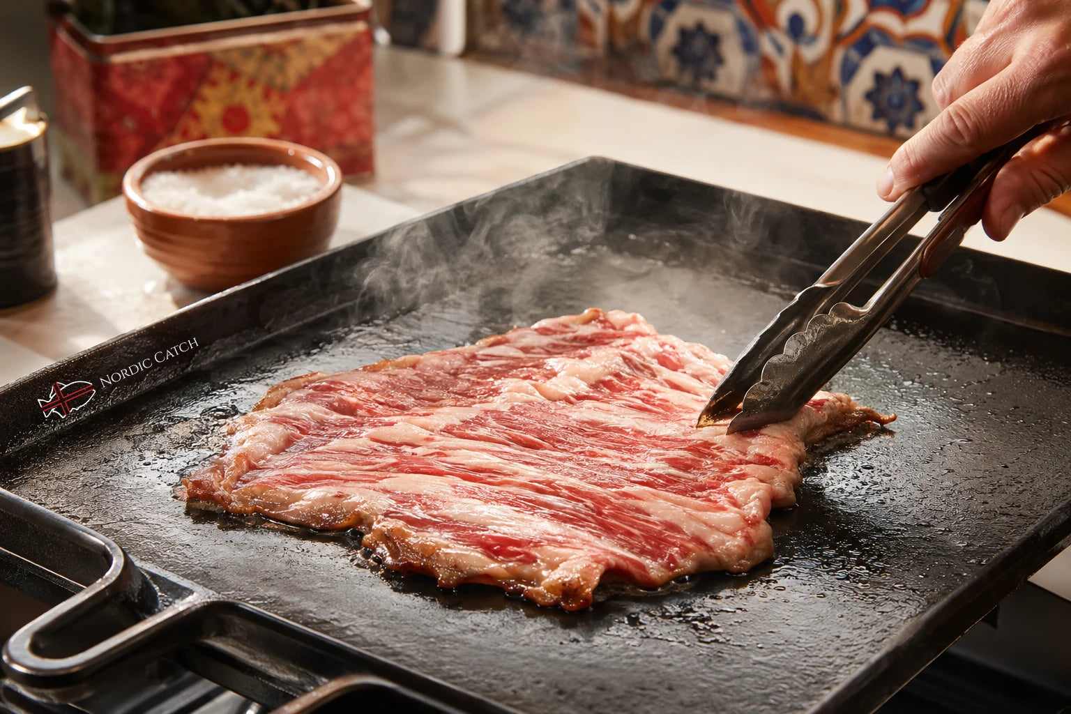 A raw marbled Iberico secreto slice cooks on a black griddle with steam rising, while a hand uses metal tongs to turn it. A small bowl of salt and a patterned container sit in the background.