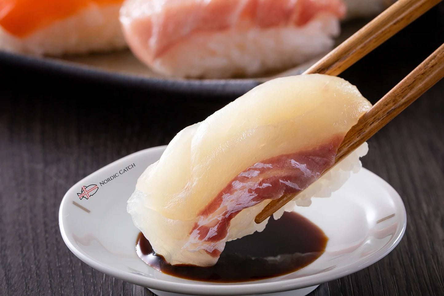 Red sea bream nigiri sushi held with wooden chopsticks over Nordic Catch branded white plate with dipping sauce and additional sushi in background