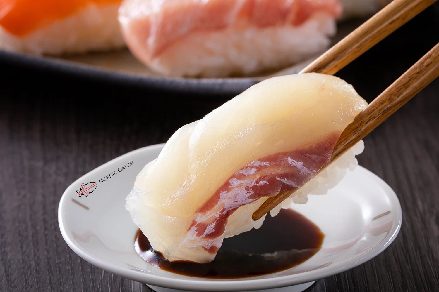 Red sea bream nigiri sushi held with wooden chopsticks over Nordic Catch branded white plate with dipping sauce and additional sushi in background
