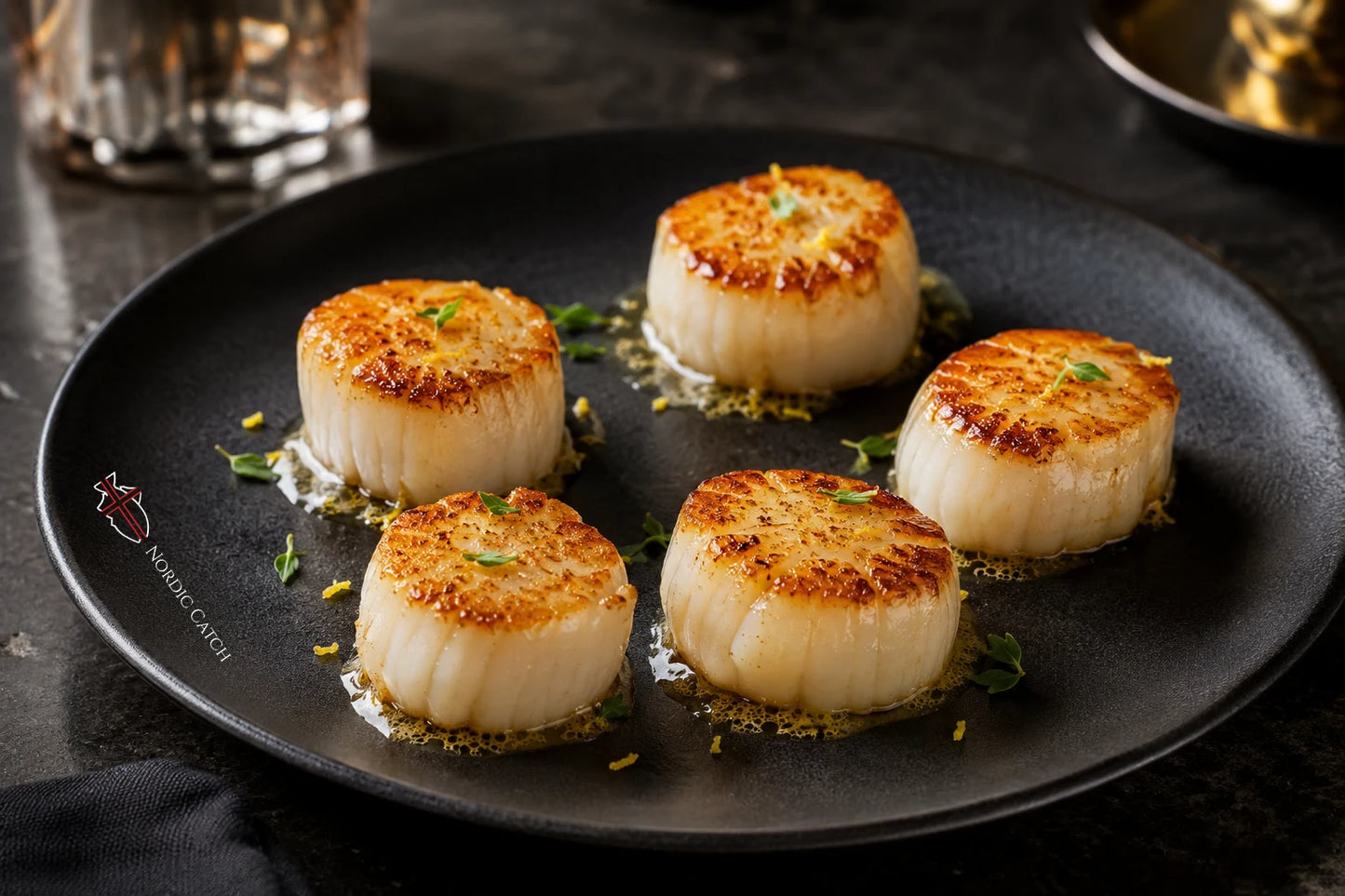 Four seared scallops on a dark plate with a small logo that reads 'Nordic Catch' on the bottom left edge of the plate, set on a dark table with a glass in the background.