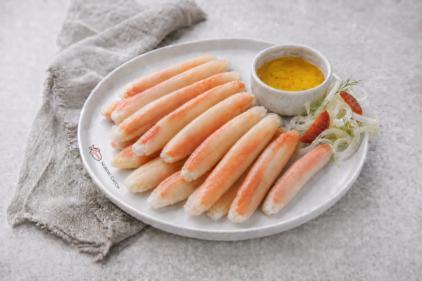 A white oval plate holding several pale orange-pink snow crab sticks, garnished with shredded white onion, a red slice, and a small cup of yellow dipping butter, set on a light gray surface with a folded beige cloth.