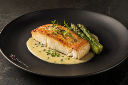 A plated cooked fish fillet with a creamy sauce, asparagus, and herb garnish on a dark plate. The plate has a visible 'NORFOLK CATCH' logo near the lower left edge.