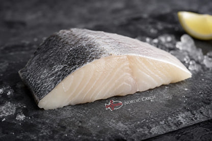A raw fish fillet with dark gray skin and a white, flaky interior, resting on a dark slate surface with ice crystals and a lemon wedge in the background. A 'Nordic Catch' logo is visible on the slate.