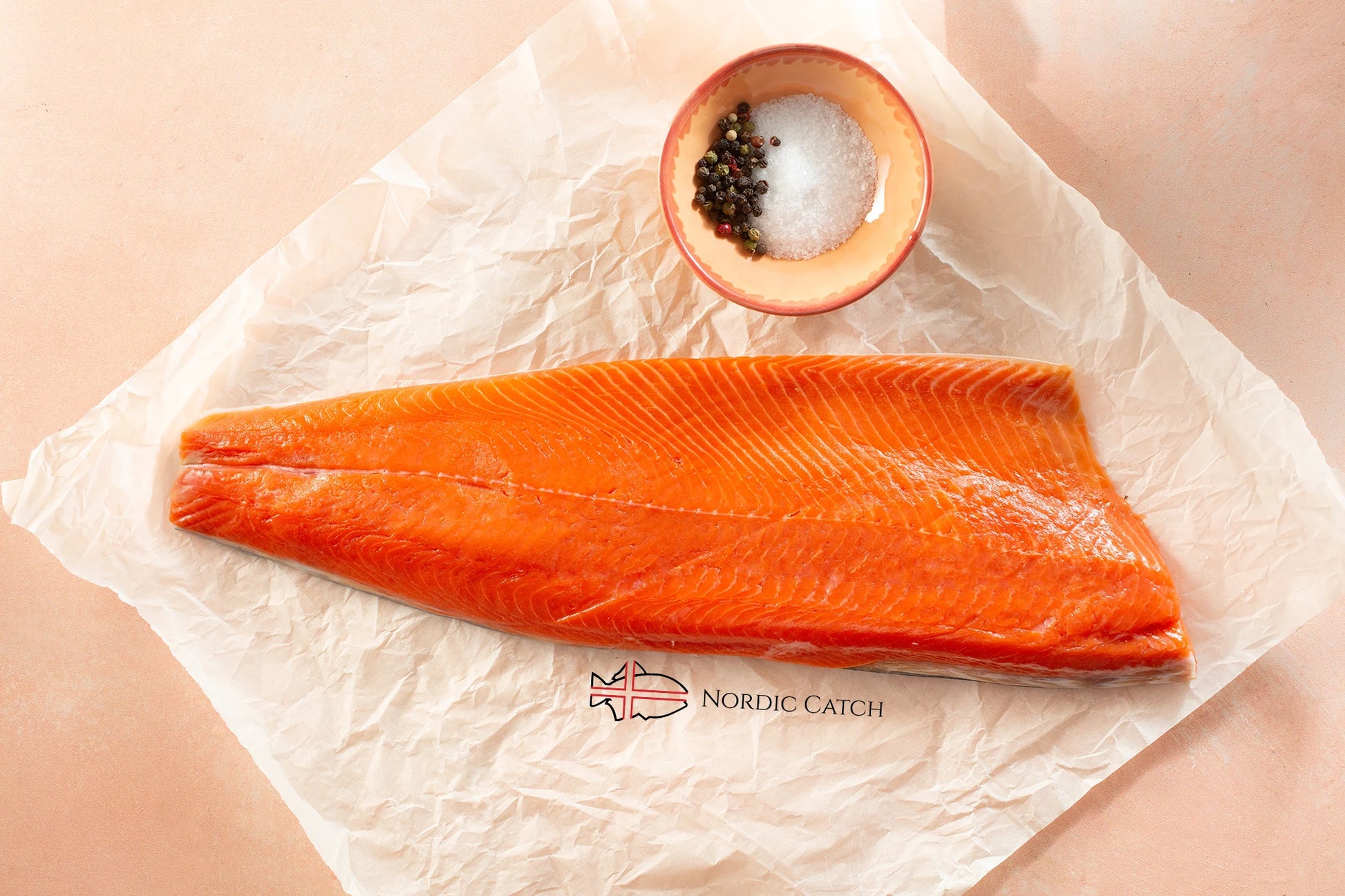 Raw salmon fillet on a piece of paper with a small bowl of salt and pepper, branded 'Nordic Catch'.