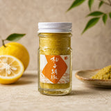 Jar of mustard with a label, lemons, and a bowl on a wooden surface